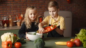 Little girl and boy mixing cutted pepper with other fresh ingredients for salad in big bowl while cooking at table with various fresh vegetables and fruits